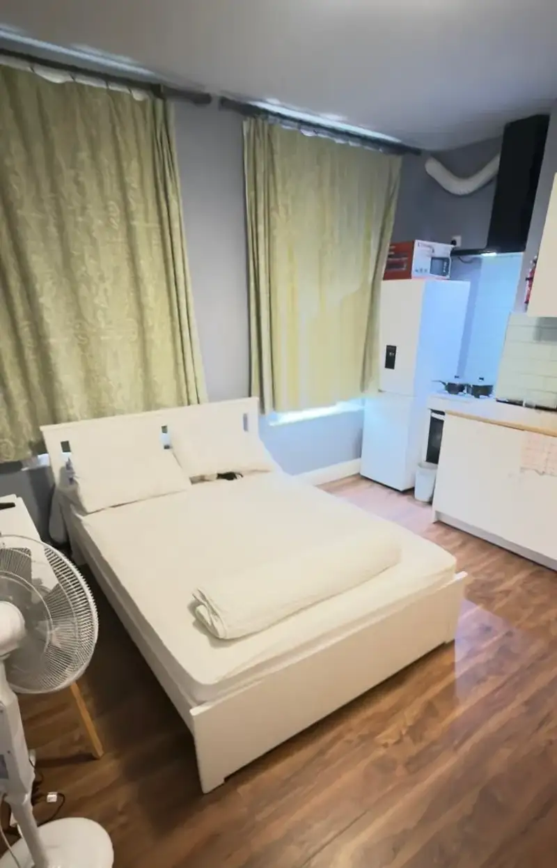 Bedroom with a double bed, pillows, folded blanket, window with drawn curtains, standing fan, and a small kitchenette with refrigerator in the corner.