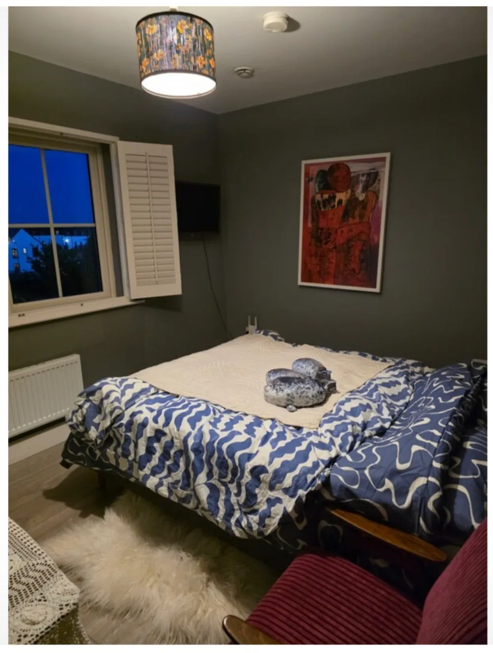 Bedroom with a double bed covered in patterned duvet, window with shutter, wall-mounted TV, framed artwork, ceiling light, and a small rug on the floor.