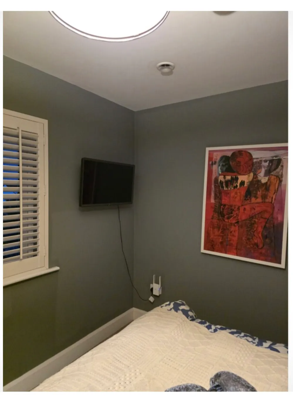 Bedroom corner with a bed in the foreground, wall-mounted TV, framed artwork, shuttered window, and a device with cables on the wall.