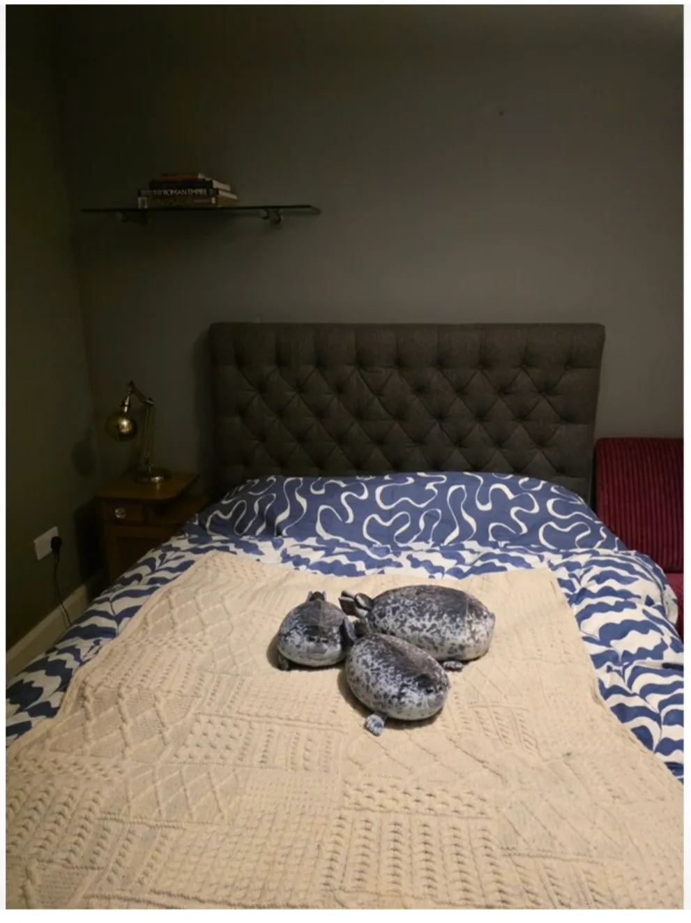 Bedroom with a double bed, tufted gray headboard, patterned blue-and-white duvet, beige knitted throw and three small plush toys on the bed; bedside table with lamp and a wall shelf with books above.