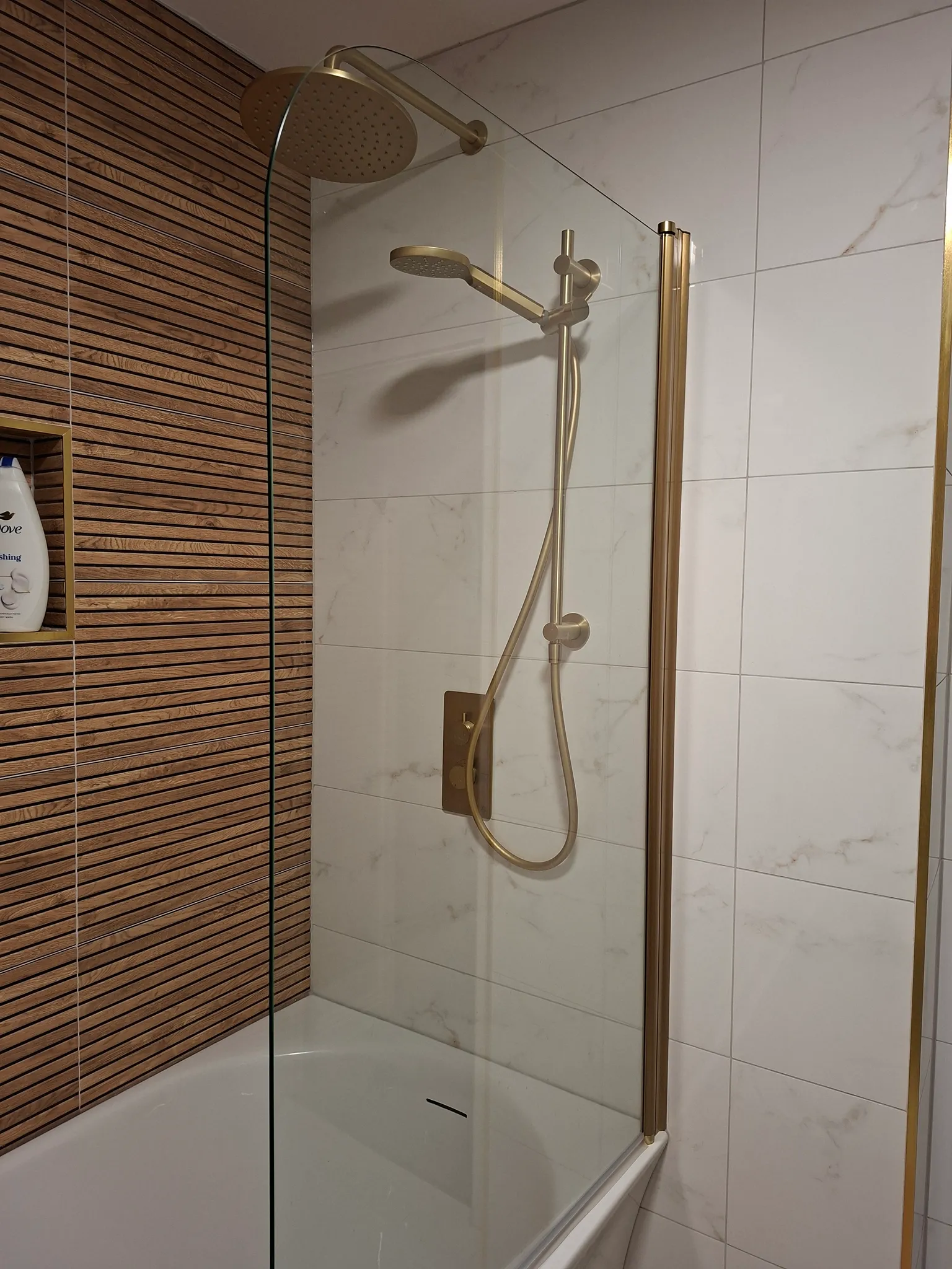 Bathroom shower enclosure with glass door, rainfall showerhead, handheld spray, and brass fixtures. Walls feature white marble-look tiles and wood-slat accent paneling on the left side.