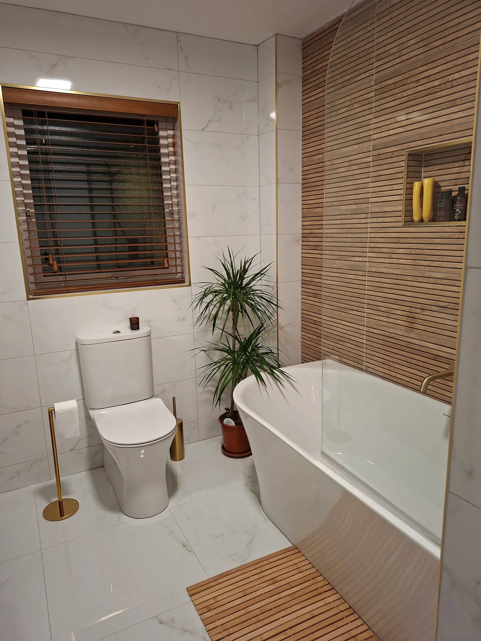 Bathroom with white toilet, bathtub with glass partition, marble-effect wall tiles, wooden slat accents, window with blinds, and potted plant.