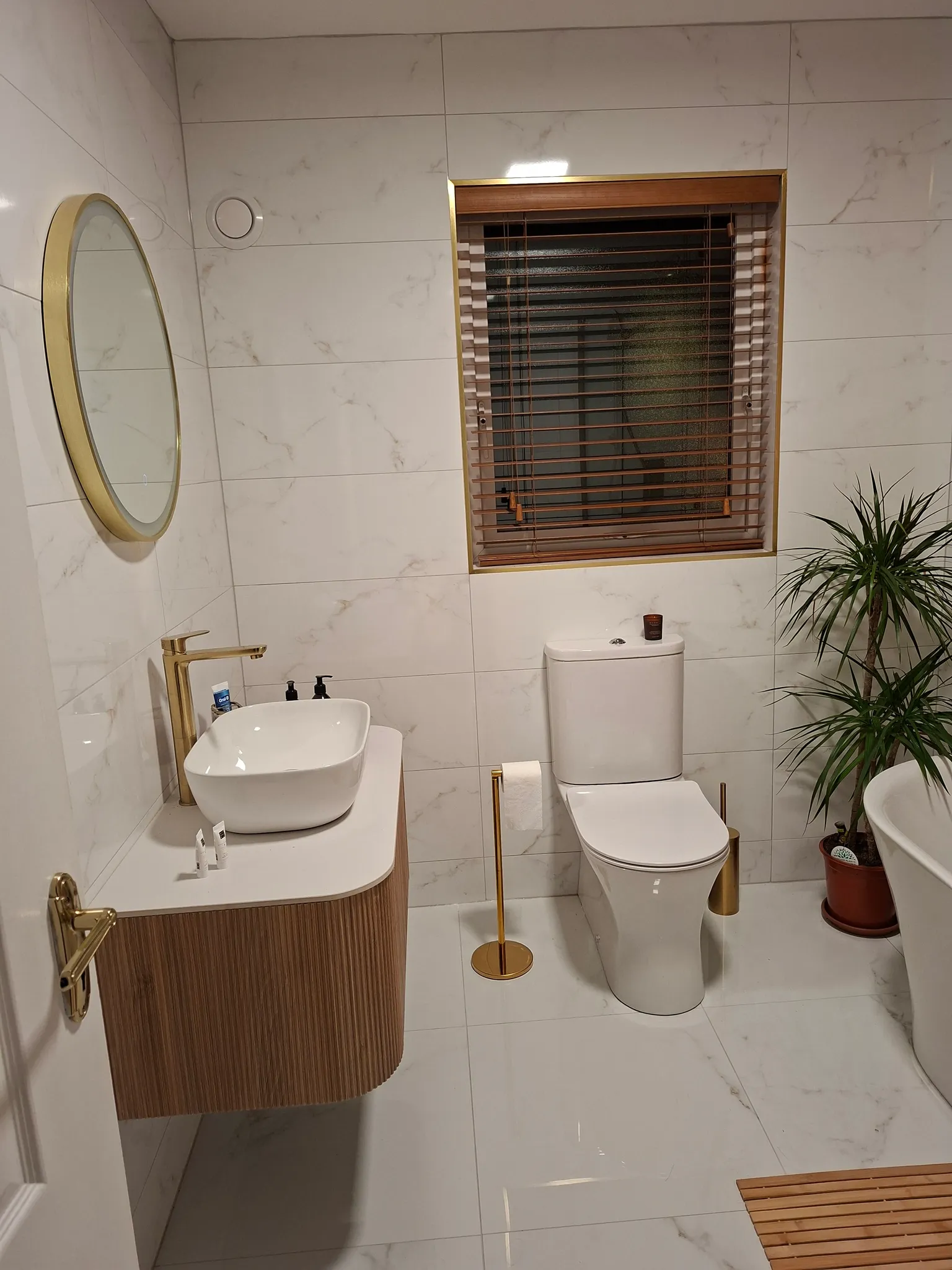 Bathroom with marble-effect wall tiles, round mirror with brass frame, white vessel sink on wooden vanity with brass faucet, toilet with brass accessories, window with wooden blinds, and potted plant.