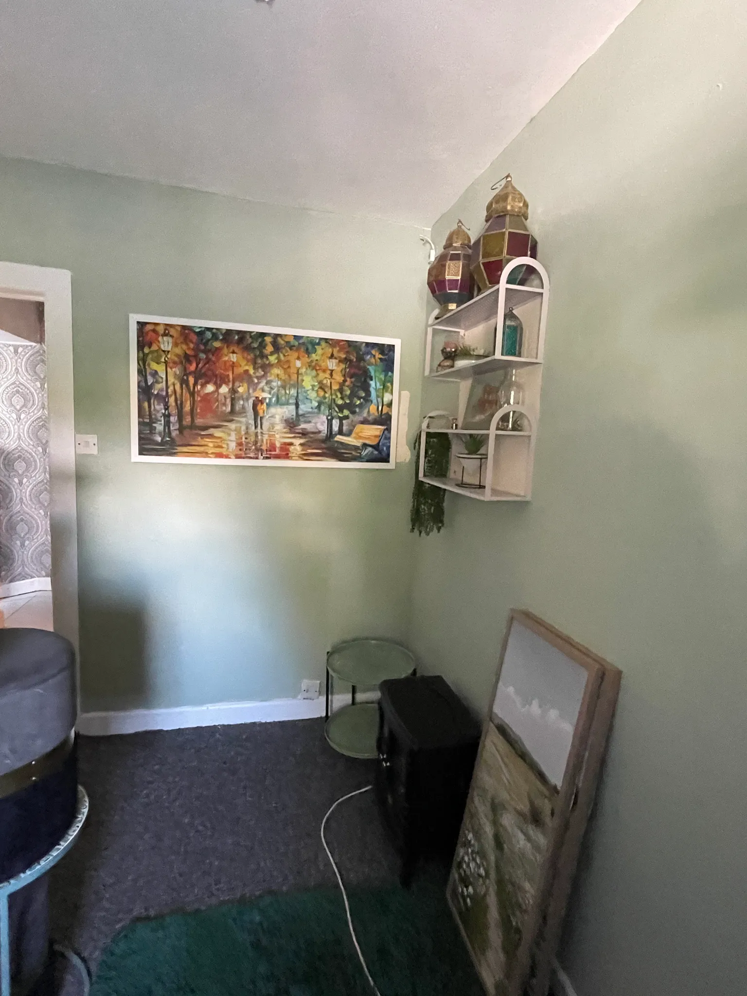 Corner of a living room with pale green walls showing a framed colorful painting, a white wall shelf with decorative lanterns and small plants, two small round nesting tables and stacked/leaning artwork on the floor.