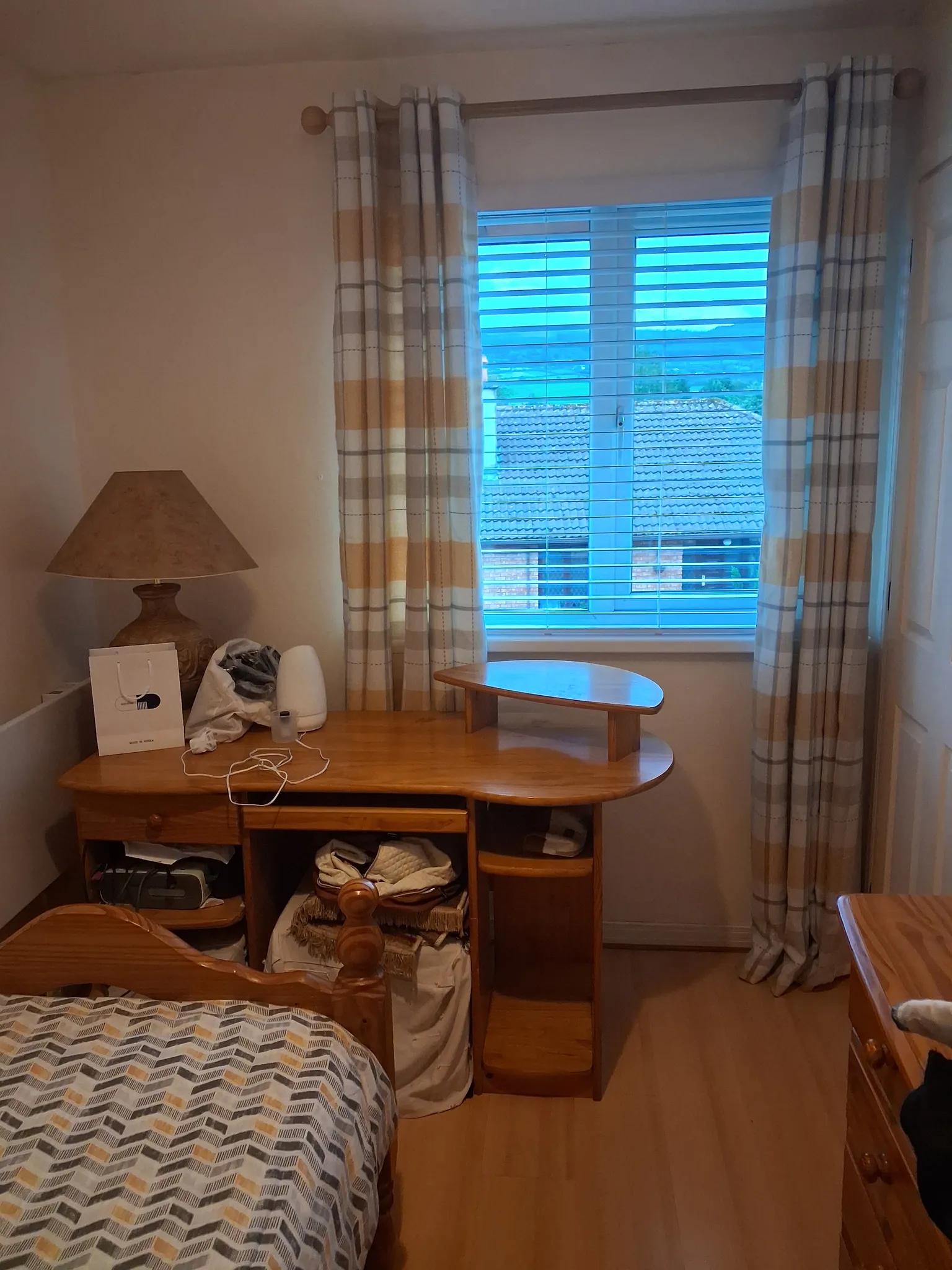 Bedroom corner with a wooden desk, lamp, patterned curtains and a window with blinds; part of a bed visible in foreground.