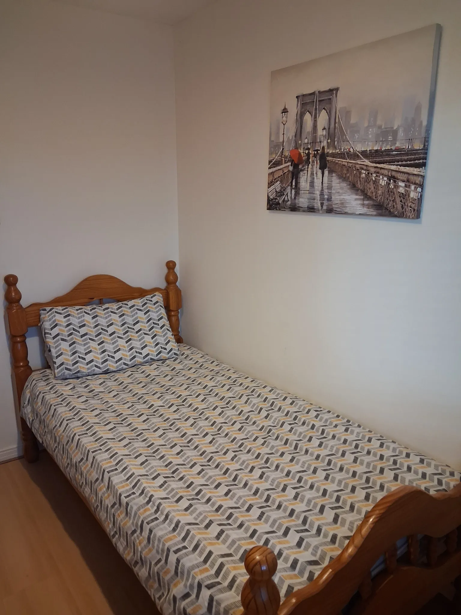 Bedroom with a single wooden bed frame, patterned bedding and pillow, and a framed bridge print on a plain wall.