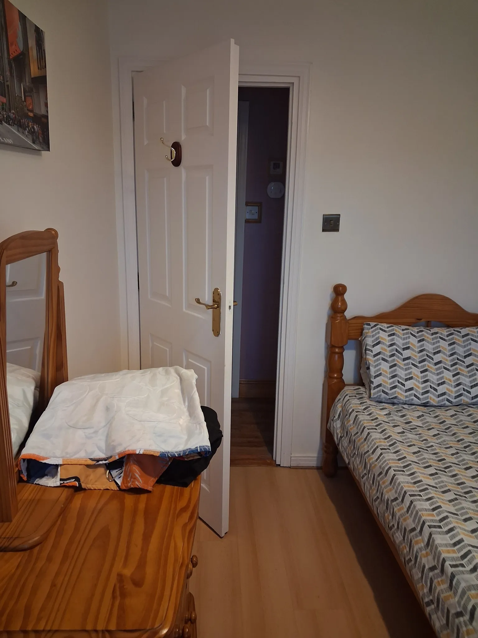 Bedroom with a single wooden bed with patterned bedding, a wooden dresser with mirror and folded linens, and a partially open white door leading to a dark hallway.