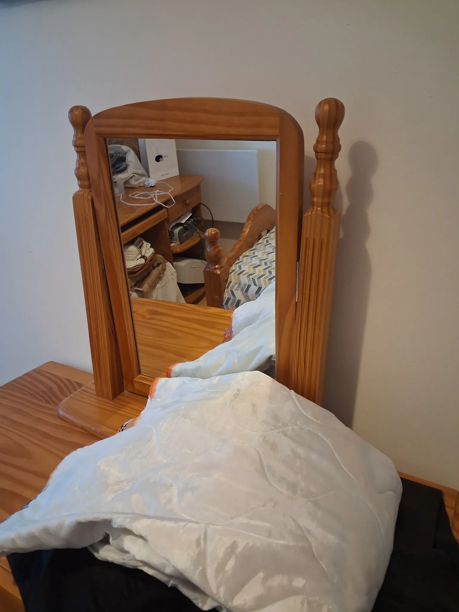 Bedroom view of a wooden framed standing mirror on a dresser with folded bedding on the surface and a reflected bedside table and headboard.