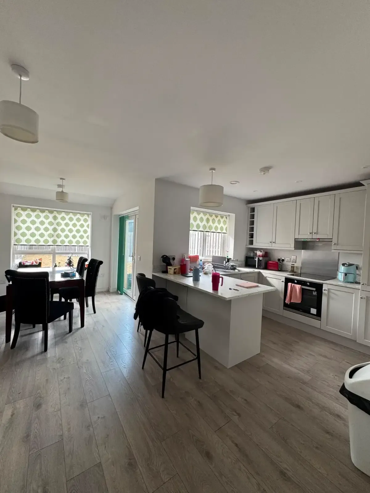 Open-plan kitchen and dining area with a central island and bar stools, dining table and chairs by two windows with patterned blinds, white cabinets and built-in oven, and light wood flooring.