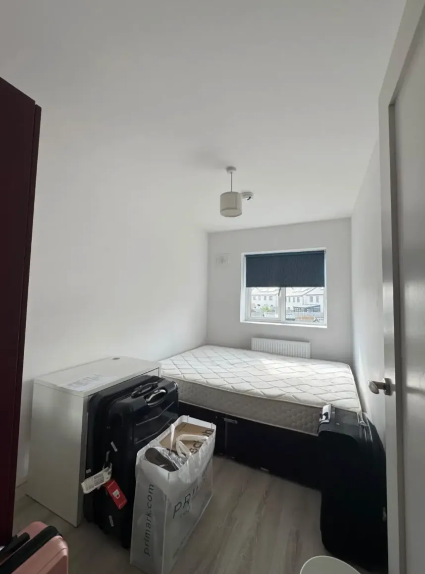 Bedroom with a mattress on a divan bed beneath a window with a closed blind; suitcases and a shopping bag on the floor.