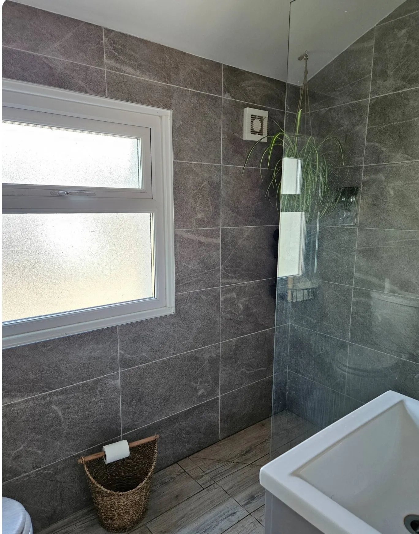 Bathroom with frosted window, grey tiled walls, glass shower screen, hanging plant, and wicker basket holding a toilet roll.