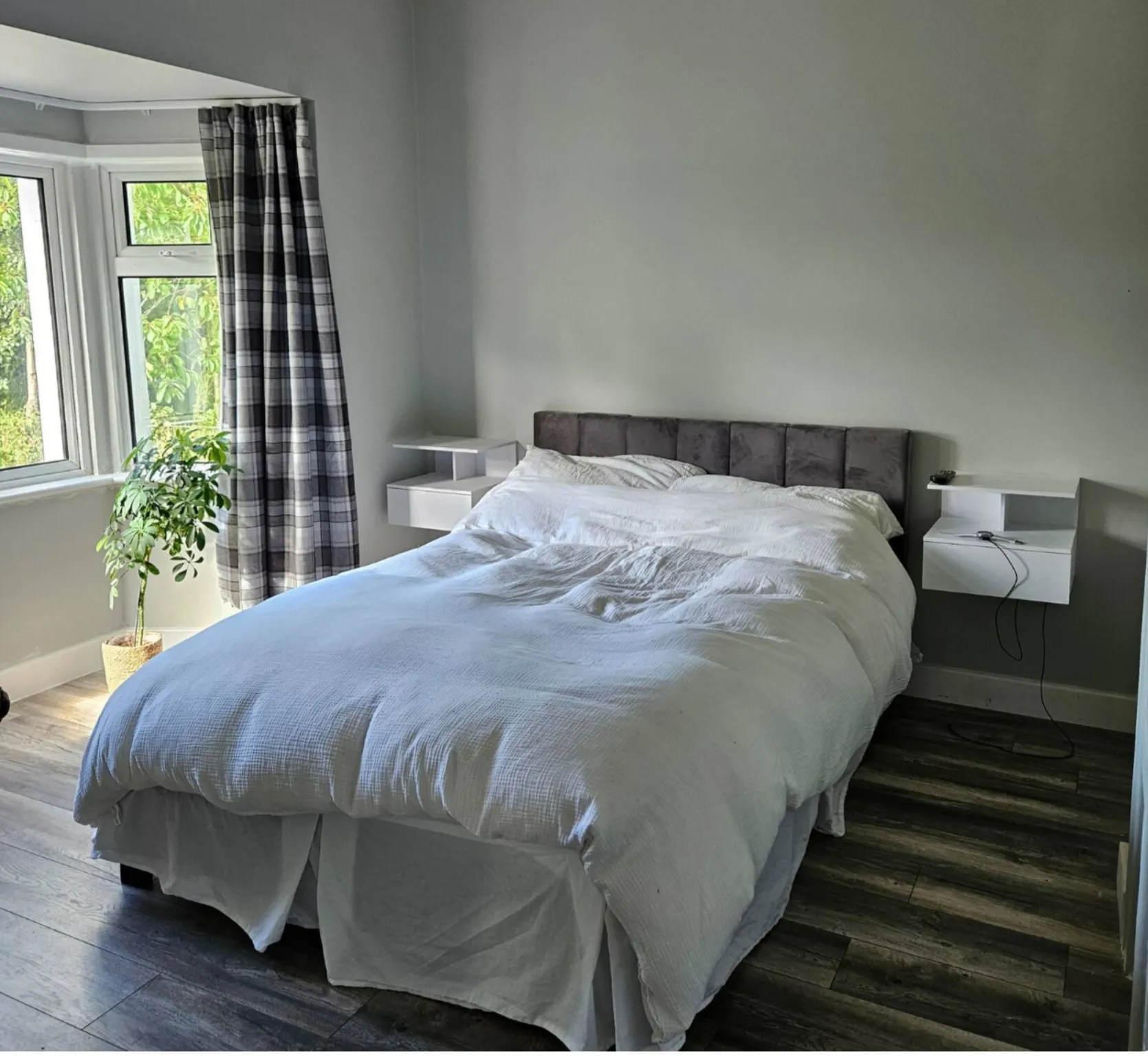 Bedroom with a double bed, white bedding, padded headboard, floating bedside shelves, window with plaid curtains and a potted plant.