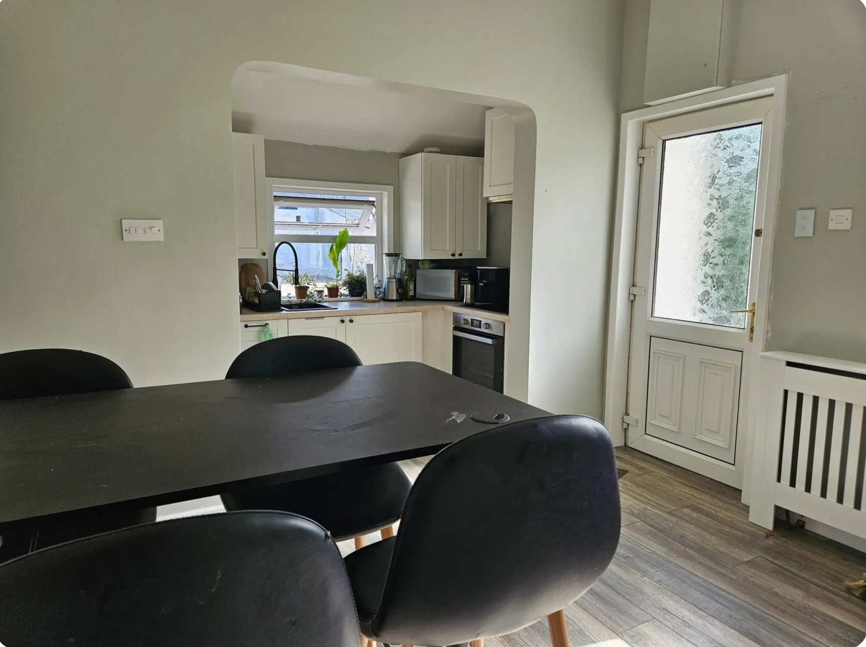 Dining area with a black dining table and chairs, open archway to a small kitchen with white cabinets, countertop appliances and a window, and a frosted glass exterior door to the right.