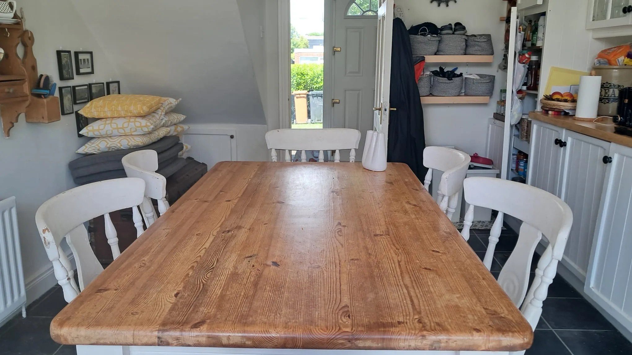Dining room with a long wooden table, white chairs, a small white vase, open door to the outside, and shelving with woven baskets and kitchen cabinets.