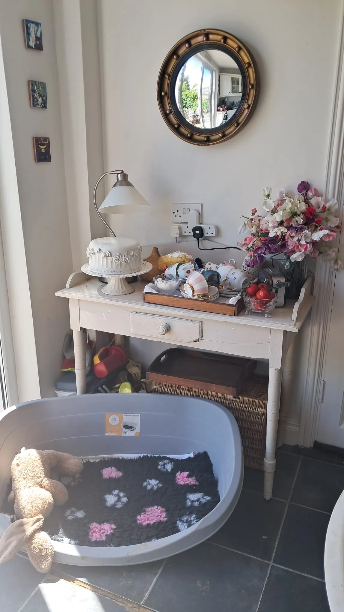 Hallway with a small white console table holding a lamp, a cake on a stand, a tray of teacups, and a vase of flowers; a round mirror hangs above and a plastic pet bed sits on the tiled floor.