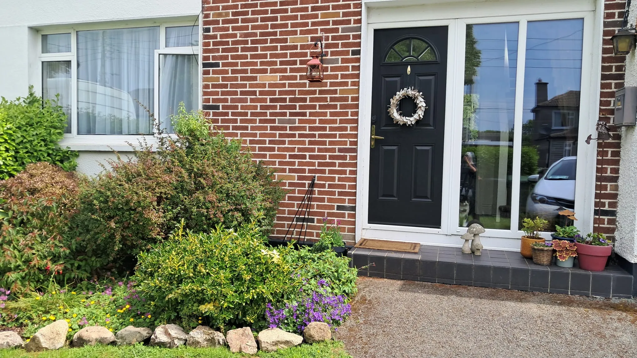 Front exterior showing a black front door with wreath, adjacent tall glass side panel, brick wall and small planted front garden with potted plants and stone edging.