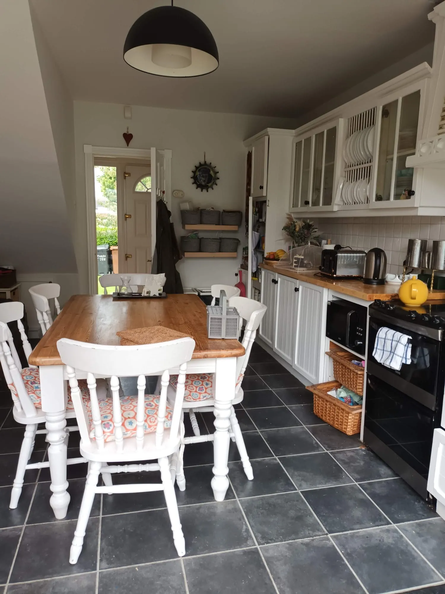 Kitchen with a wooden dining table surrounded by white chairs, white cabinets with glass doors, countertop appliances (toaster and kettle), and a door at the back.