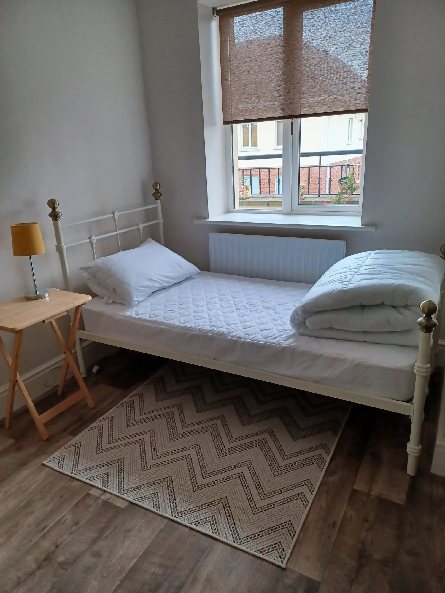 Small bedroom with a single bed, metal bed frame, pillow and folded duvet, window with a roller blind, bedside table with a lamp and a patterned rug on a wood-look floor.
