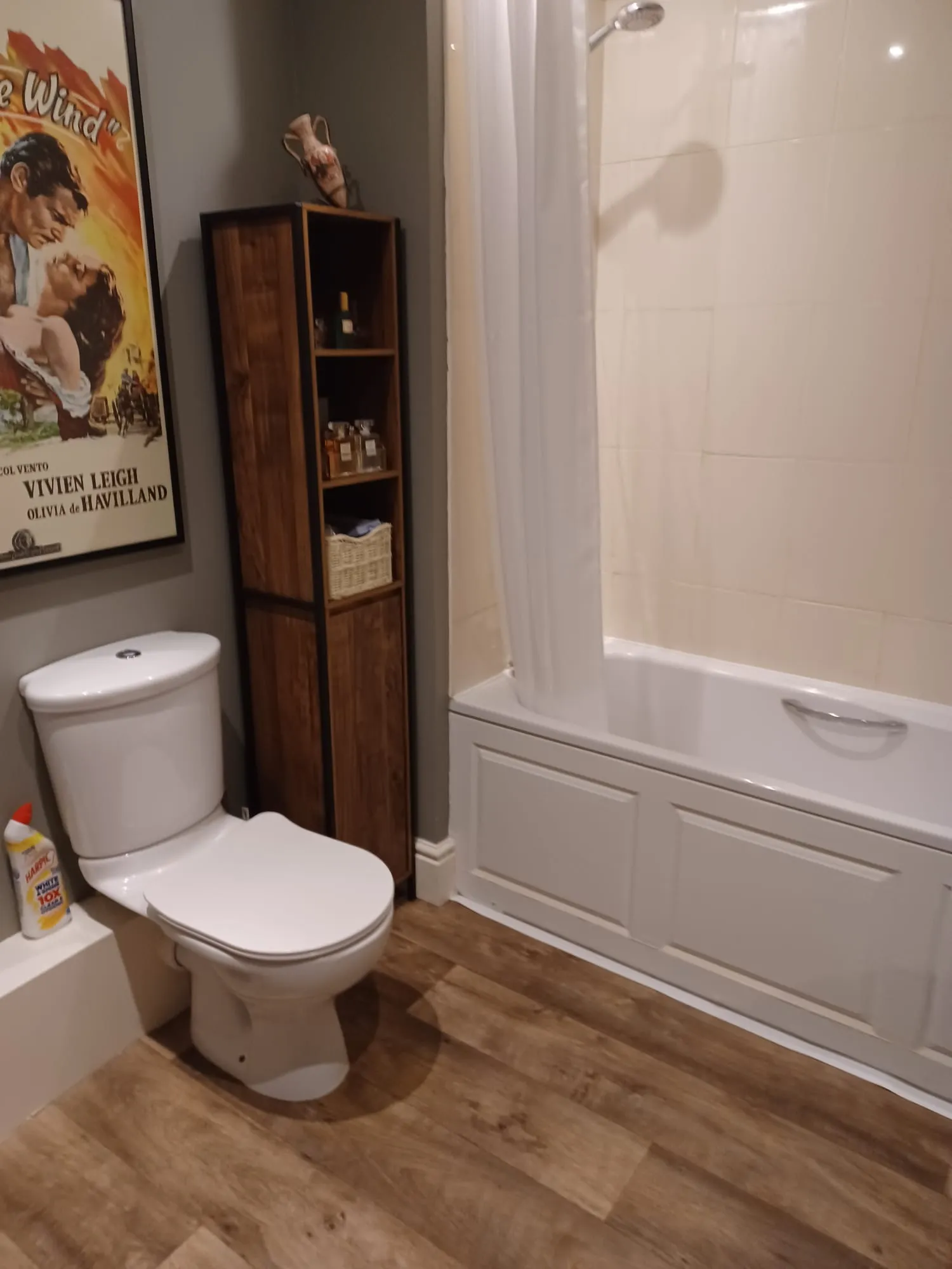 Bathroom with a toilet, bathtub and shower with a white curtain, and a tall wooden shelving unit beside the tub.