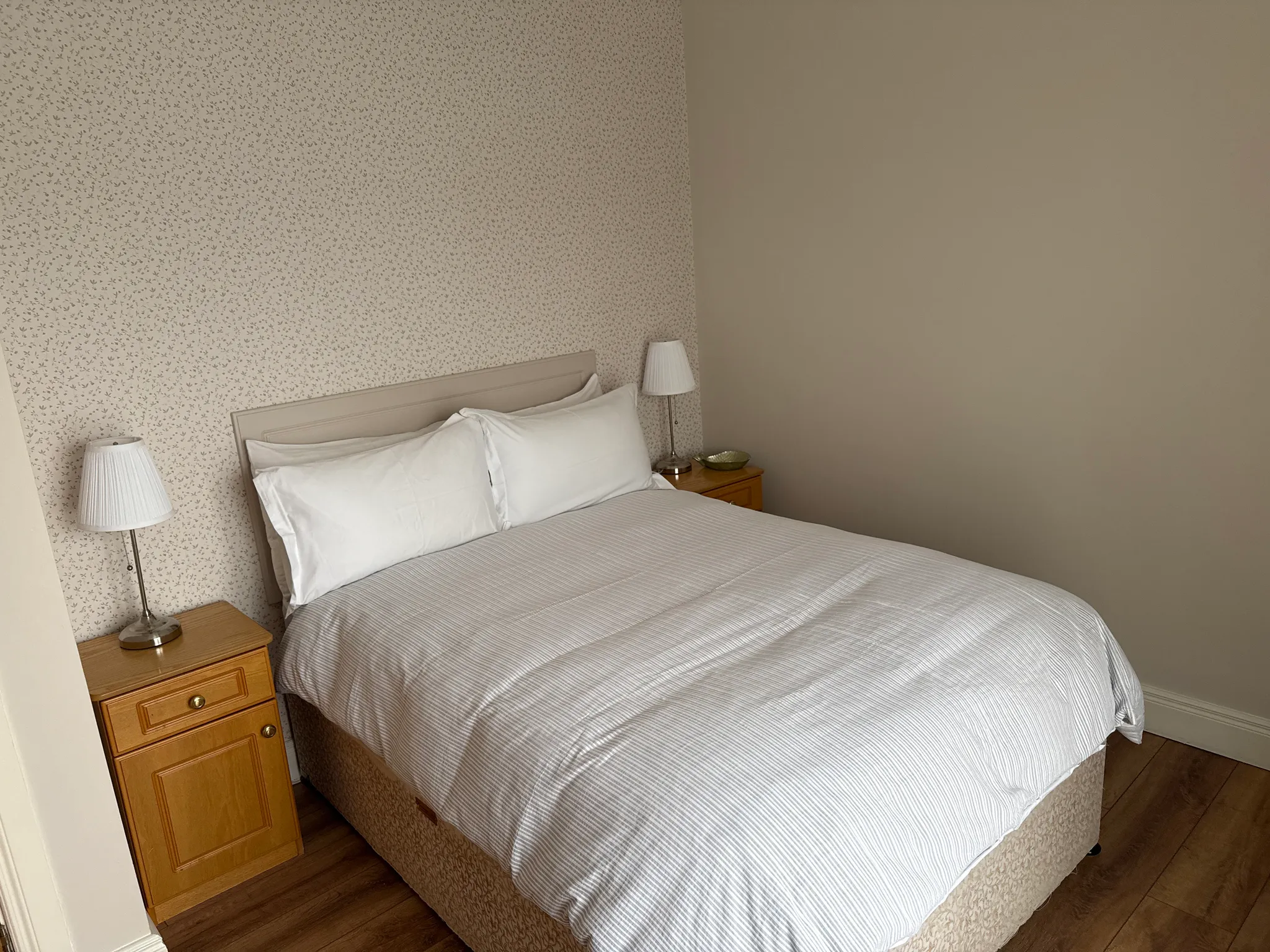 Bedroom with a double bed, striped gray and white bedding, white pillows, wooden nightstands with table lamps on either side, and patterned wallpaper