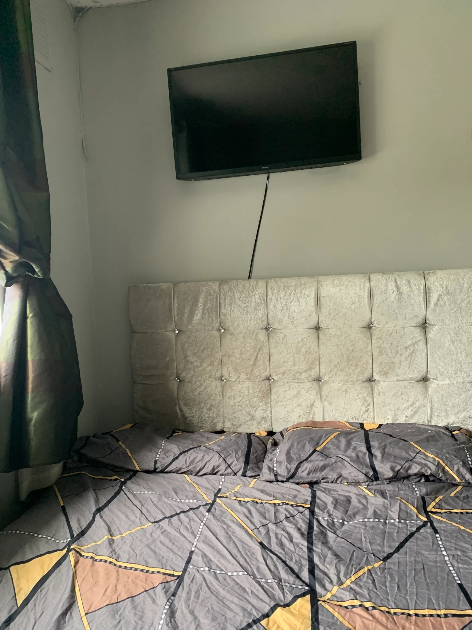 Bedroom showing a bed with a tufted headboard, patterned bedding, a wall-mounted TV above the headboard, and a curtain at the left.