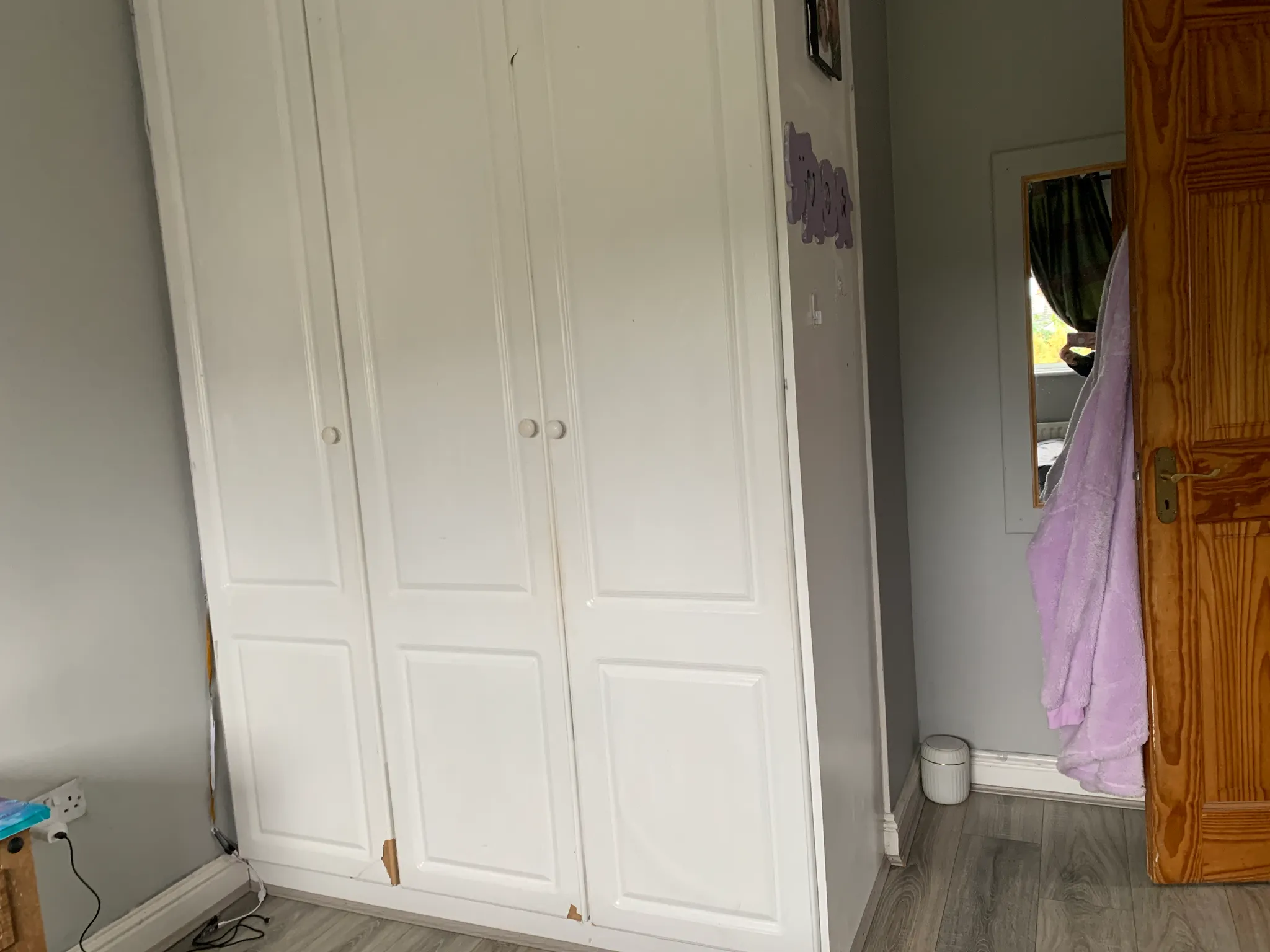 Bedroom corner showing a white built-in wardrobe with closed doors, a wooden room door with a hanging purple robe, a narrow mirror on the wall and a small trash bin on the floor.
