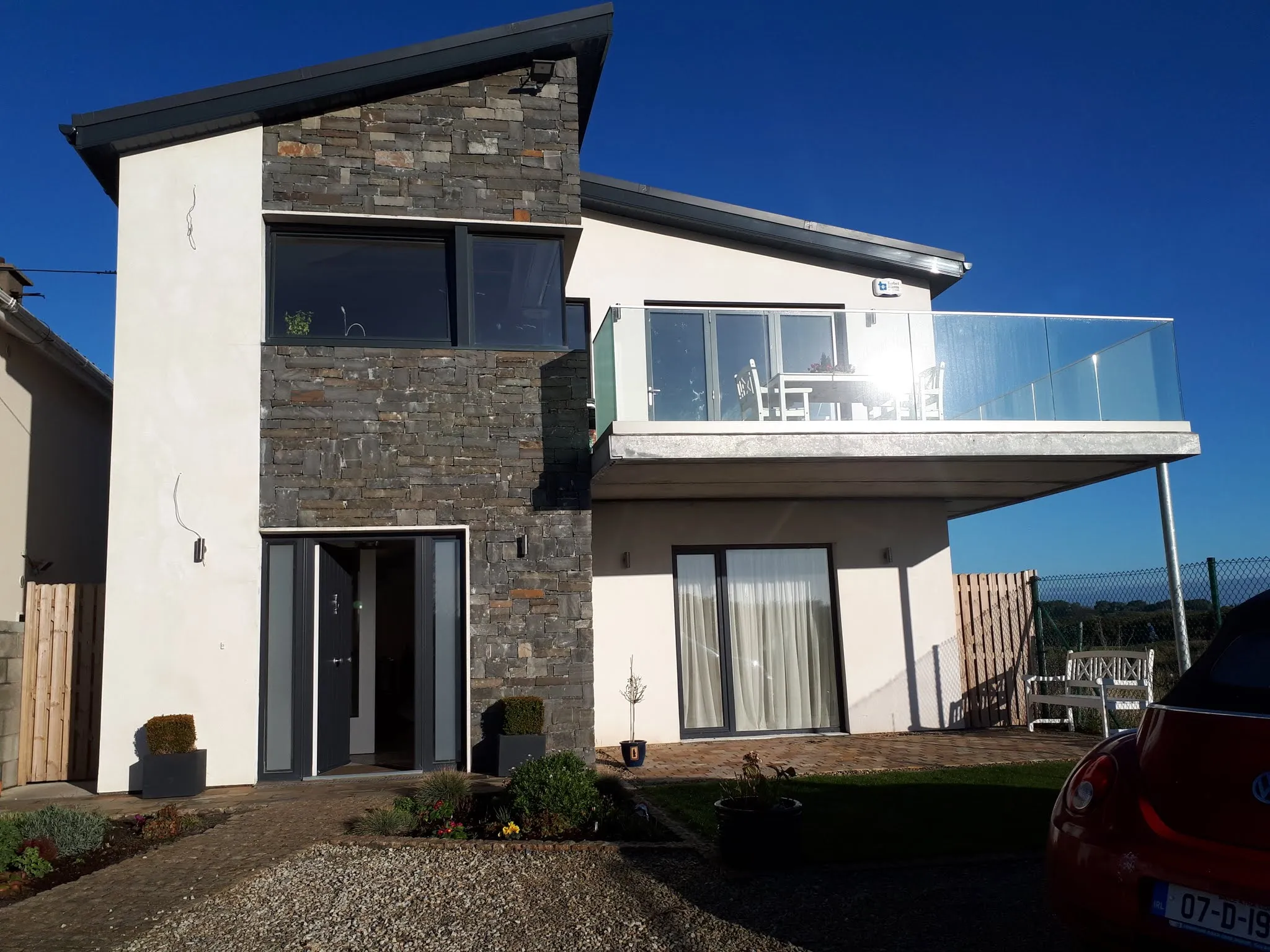 Exterior view of a two-story house with stone cladding, a glass-railed balcony, large windows and a front entrance.
