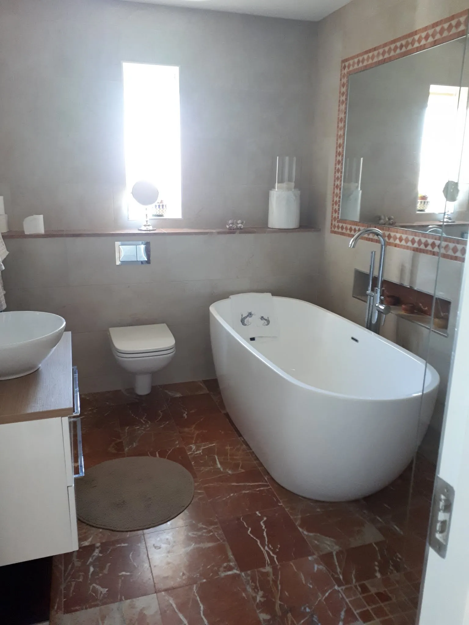 Bathroom with a freestanding bathtub, wall-mounted toilet, countertop sink, large wall mirror and a narrow vertical window.