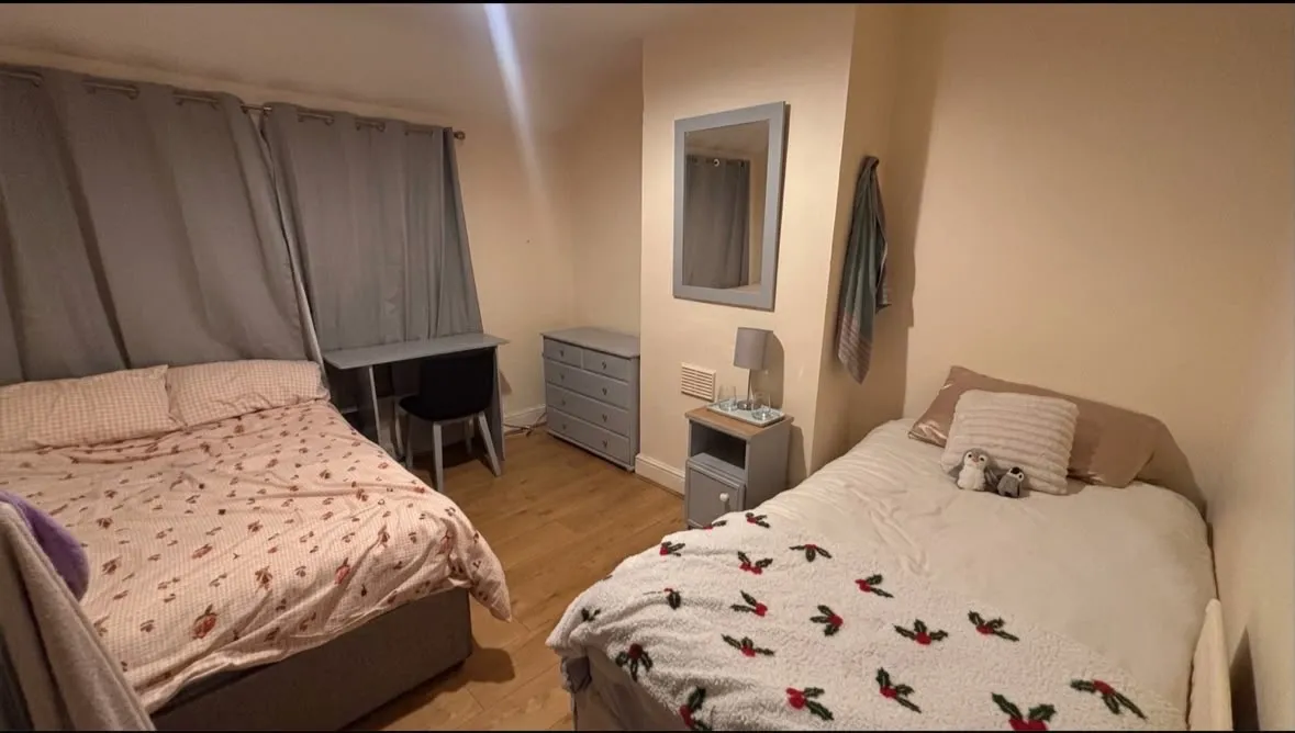 Bedroom with two single/double beds, a desk and chair by a curtained window, a chest of drawers, bedside table with lamp and wall mirror.