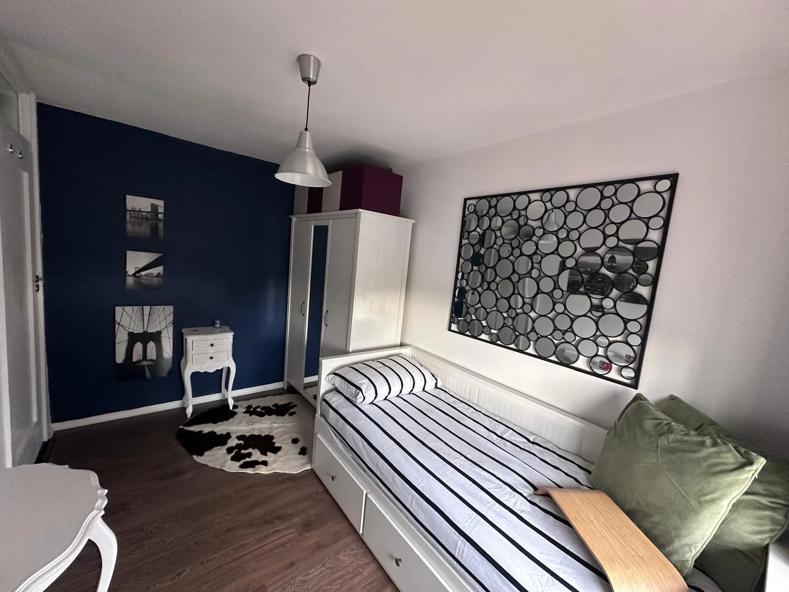 Bedroom with a single bed with striped bedding, wardrobe, small bedside table, decorative circular mirror panel on the wall, and pendant ceiling light.