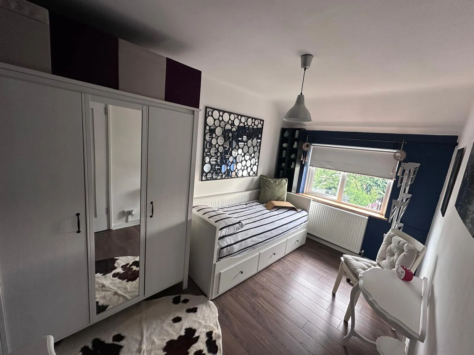 Bedroom with a single daybed with striped bedding and storage drawers, a wardrobe with mirrored door, window with a roller blind, a cushioned chair and small round table, pendant light and wall mirror art.