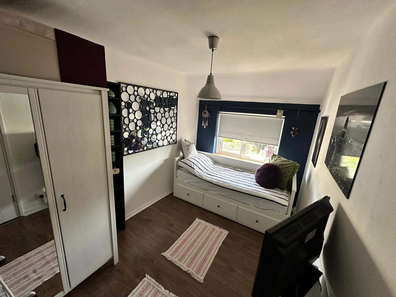 Bedroom with a single daybed under a window with a roller blind, white wardrobe with mirrored door, circular mirror wall art, pendant ceiling light and hardwood floor.