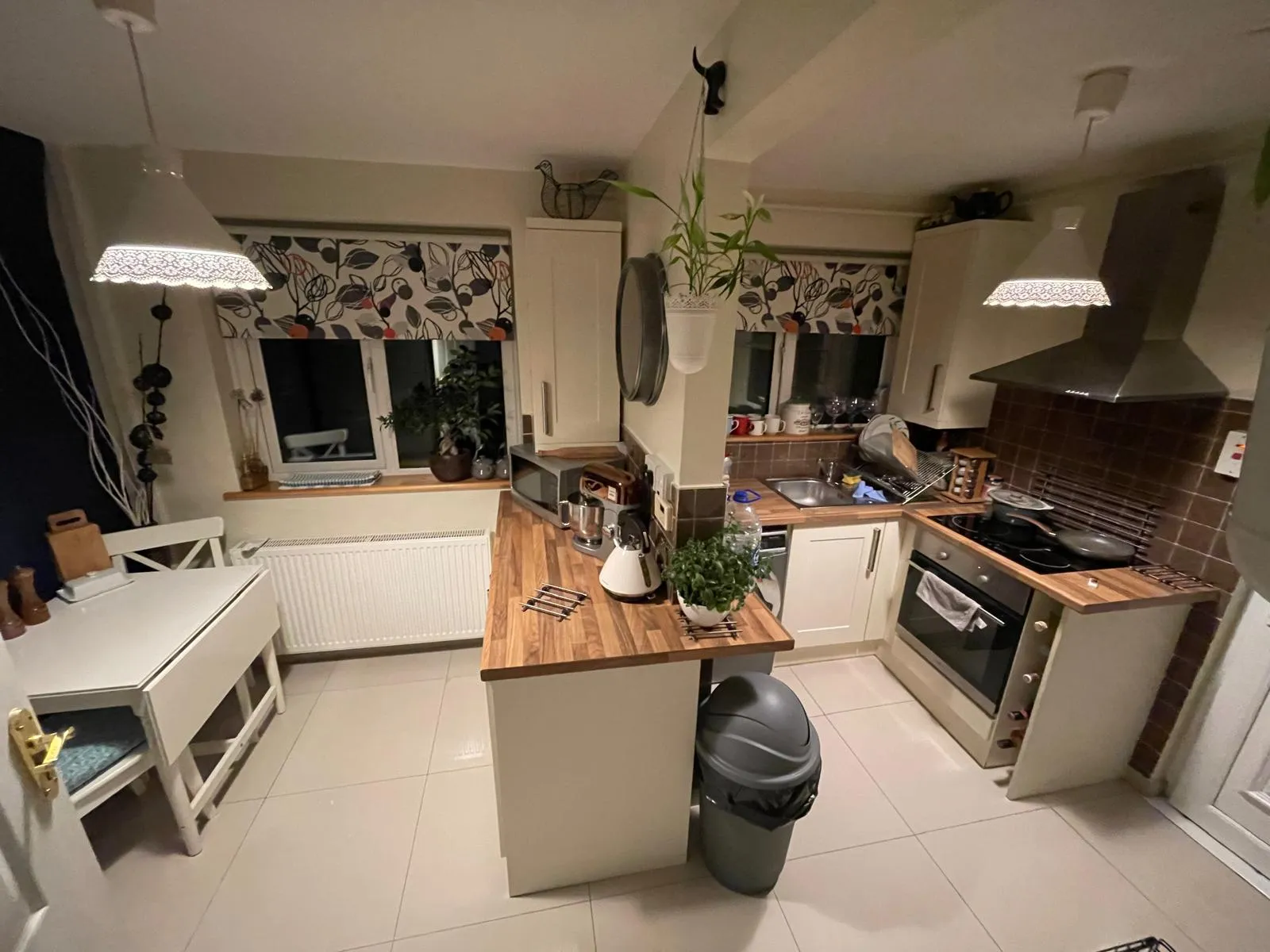 Kitchen with a central island and wooden countertops, built-in oven and stovetop, sink by a window with patterned blinds, pendant lights, hanging plant and a trash bin.