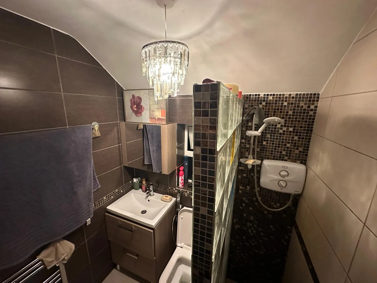 Small bathroom with a sink and vanity, toilet, glass block partition leading to a shower with handheld showerhead, mosaic tile wall, towel on a rack, and a hanging chandelier light.