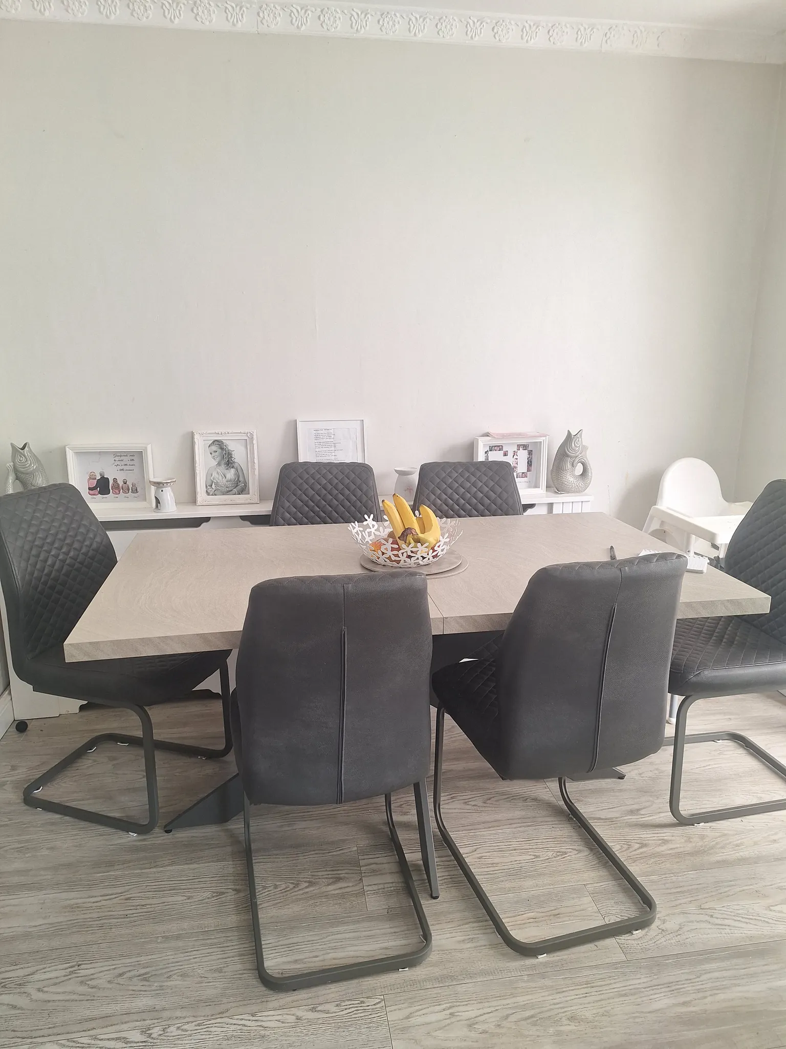 Dining room with a rectangular table, six dark upholstered chairs, a decorative fruit bowl on the table and framed photos on a low shelf against a plain wall.