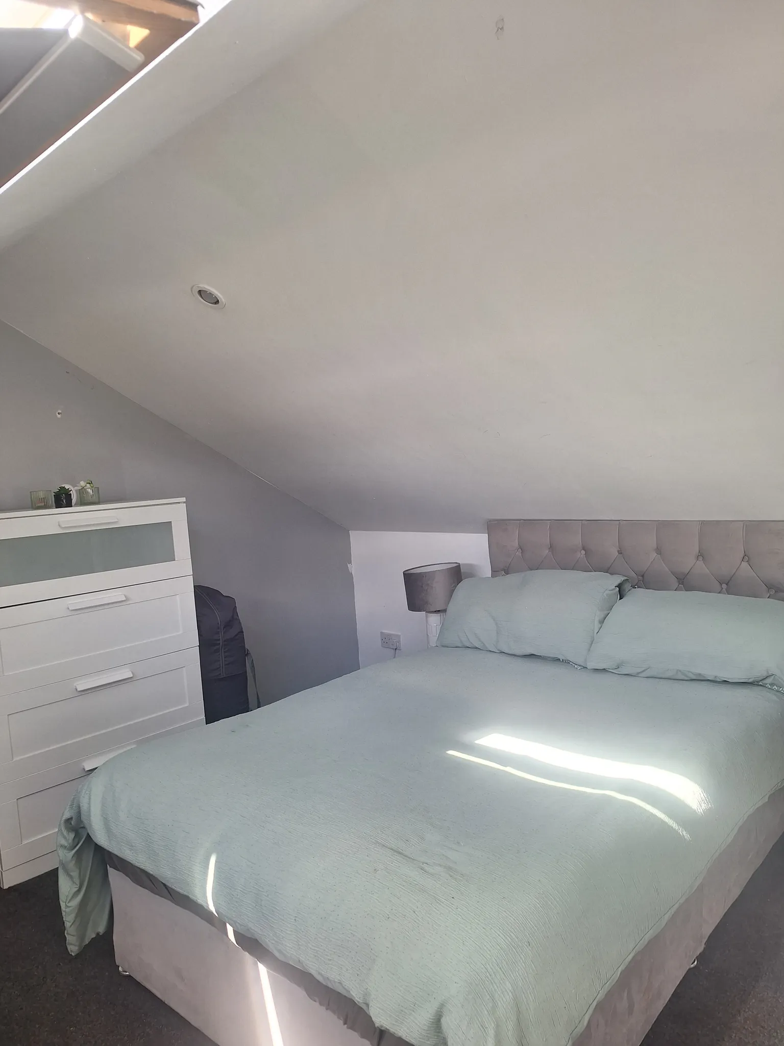 Bedroom with a double bed and tufted headboard under a sloped ceiling, bedside table with lamp and a white dresser visible