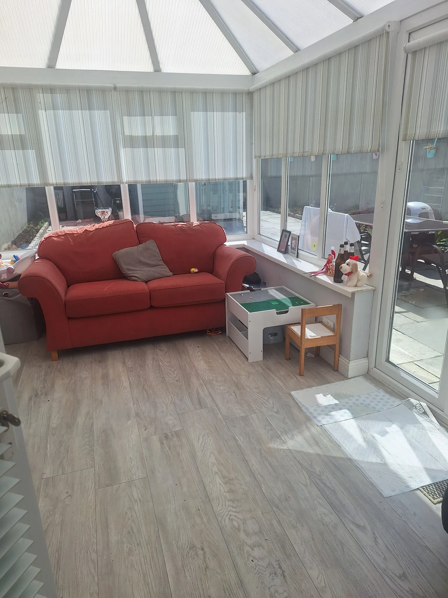 Conservatory/living room with a red two-seat sofa, striped roller blinds on multiple windows, a small children's play table and wooden floor.