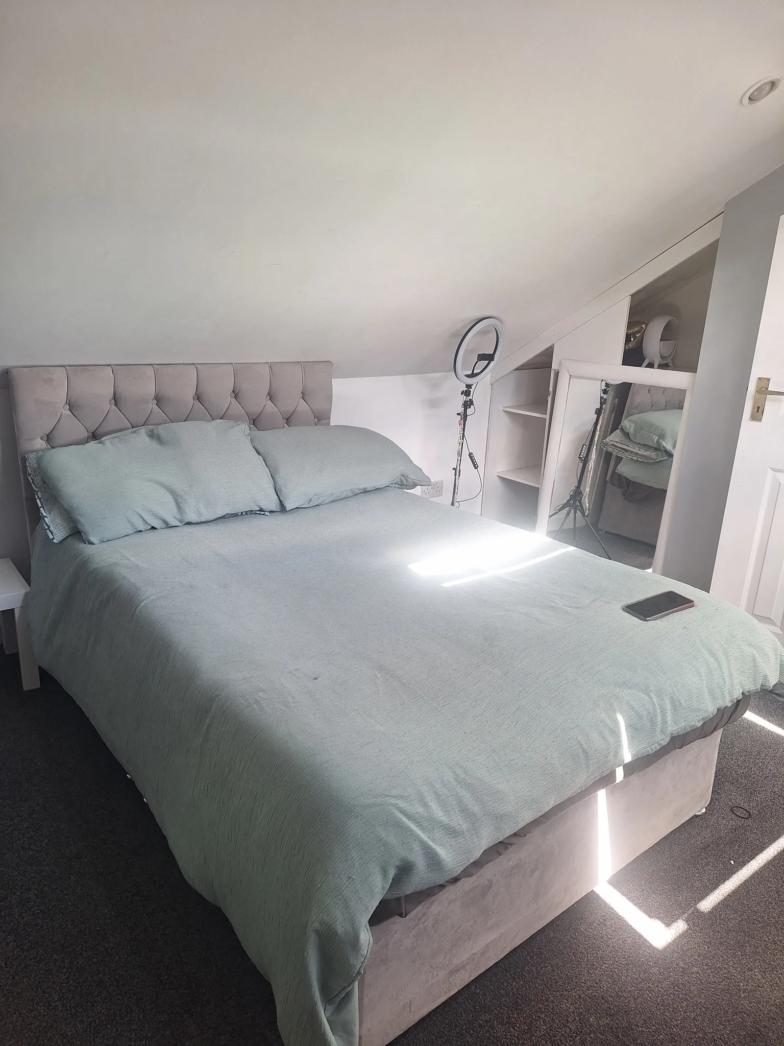 Bedroom with a double bed and upholstered headboard under a sloped ceiling; bedside table, ring light on a tripod, and a leaning mirror visible at the side.