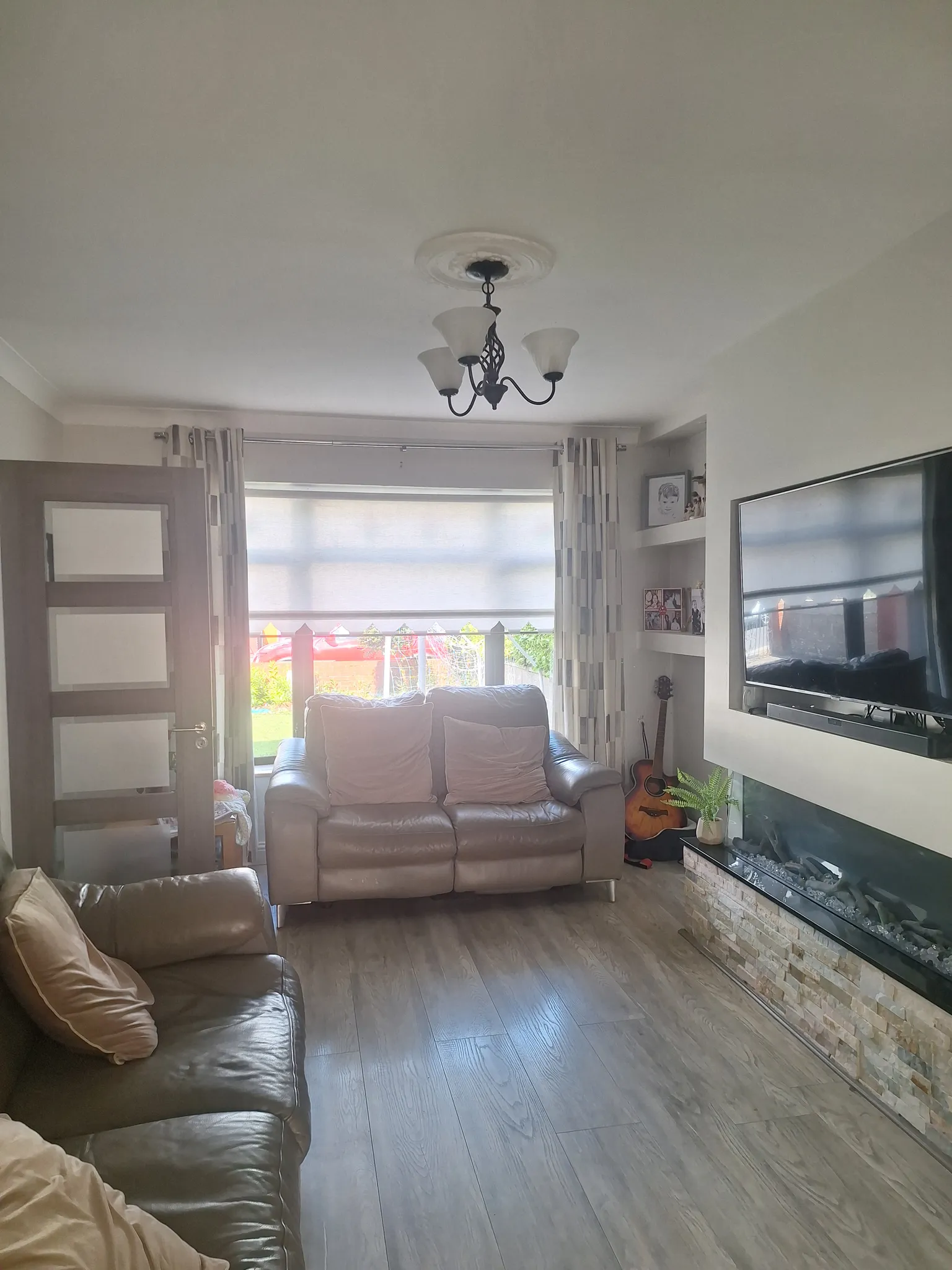 Living room with two-seat sofa and armchair facing a wall-mounted TV, window with blinds and curtains, ceiling light fixture and built-in fireplace.