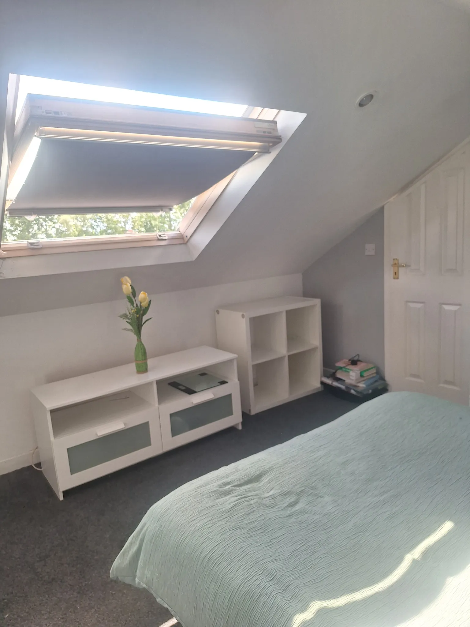 Bedroom with a skylight window and lowered blind, bed with light green coverlet, white storage units and a vase with yellow flowers.