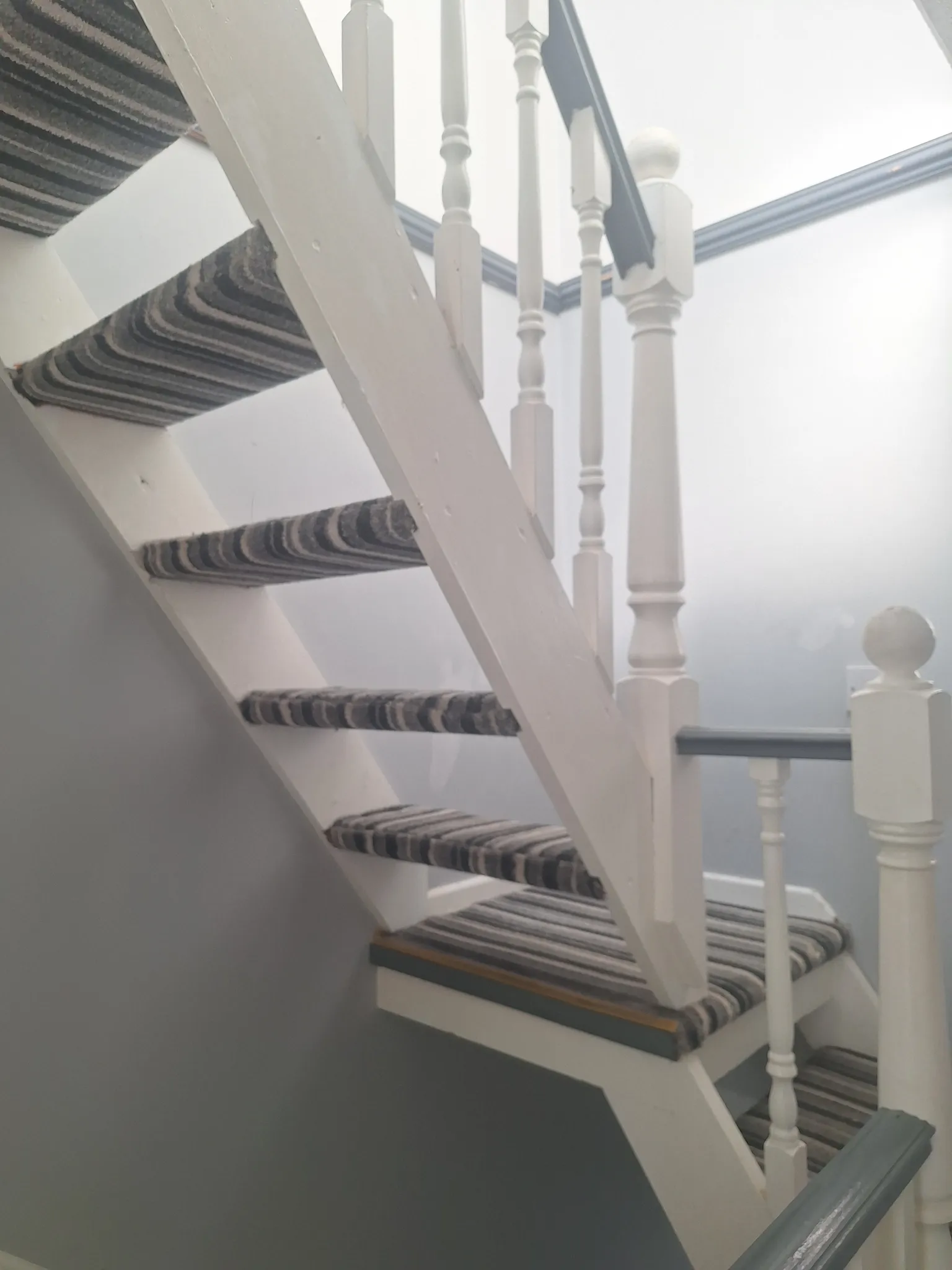 Interior staircase with white painted stringers and spindles, striped carpet treads and a grey handrail against light-coloured walls.