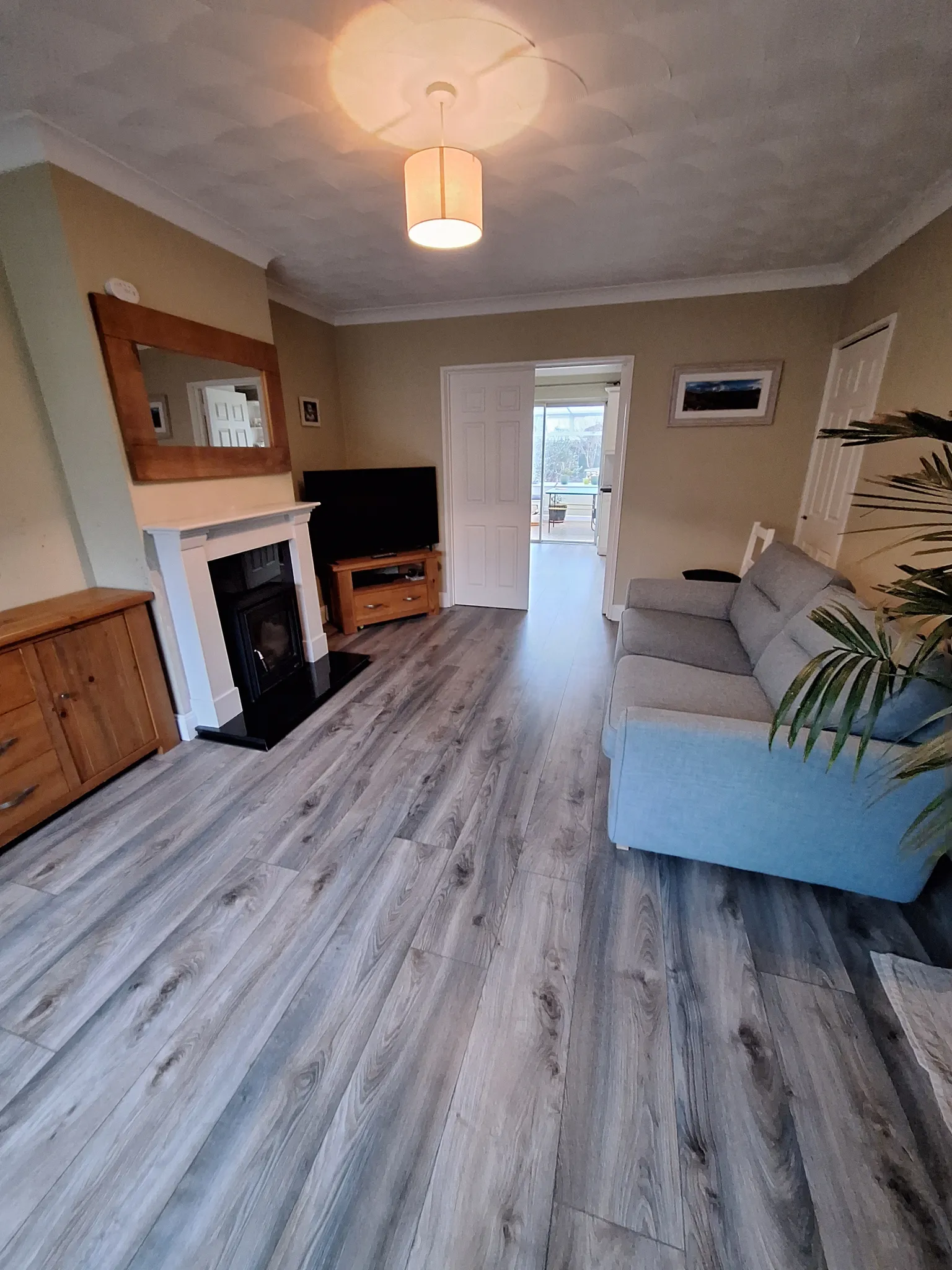 Living room with a grey sofa on the right, TV and fireplace on the left, wood-look flooring and a doorway leading to another room.