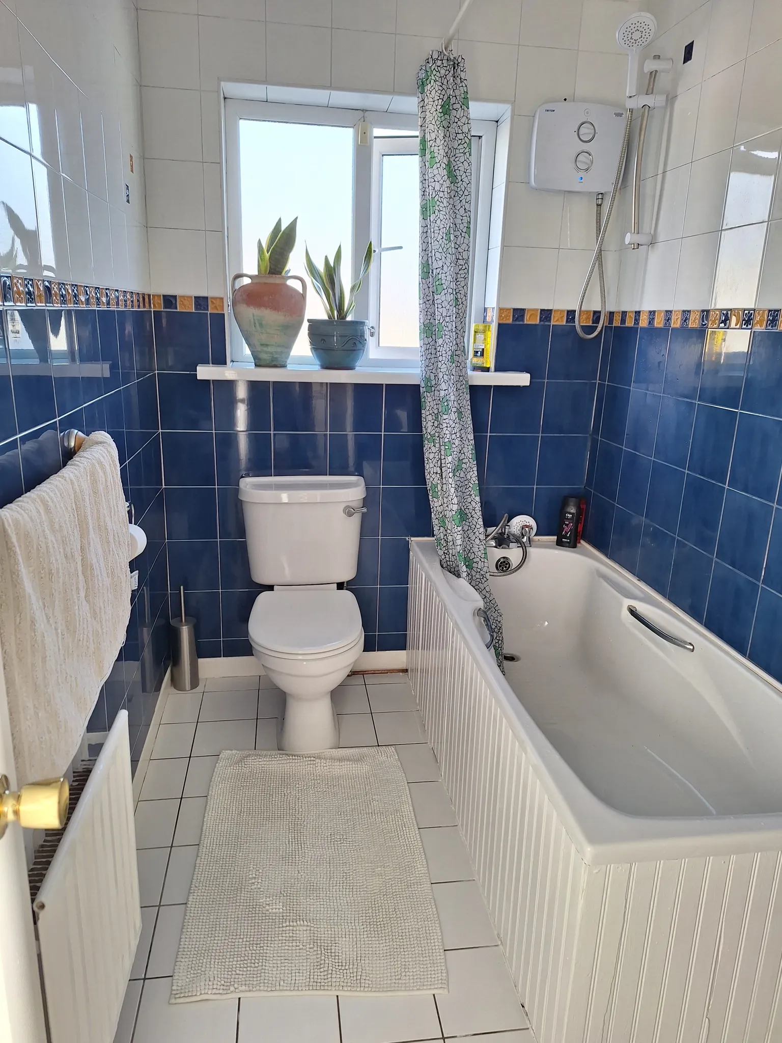 Bathroom with a toilet, bathtub with showerhead and curtain, tiled walls and a window with two potted plants on the sill.