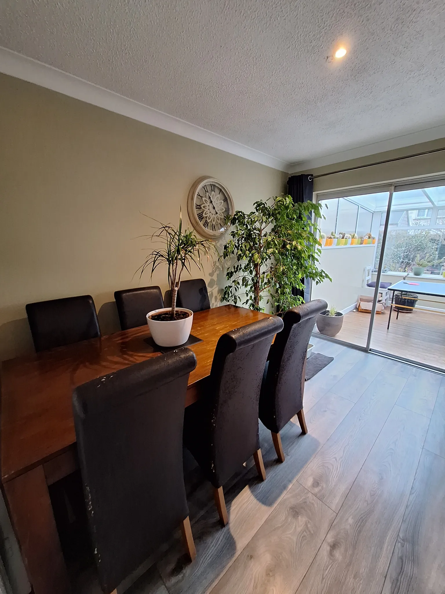 Dining room with a wooden table, six dark chairs, potted plants, a round wall clock, and sliding glass door to a glazed sunroom.