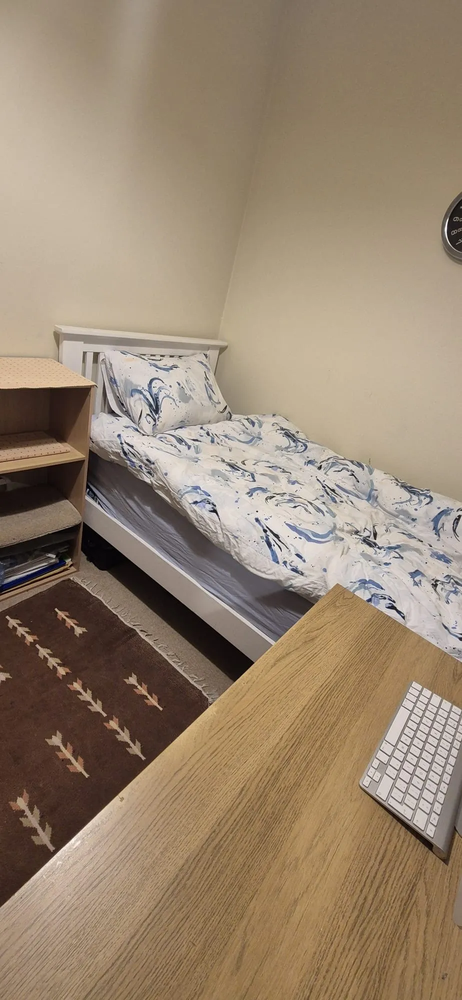 Small bedroom with a single bed against a corner, patterned duvet and pillow, wooden desk with keyboard in foreground, small shelving unit and rug.