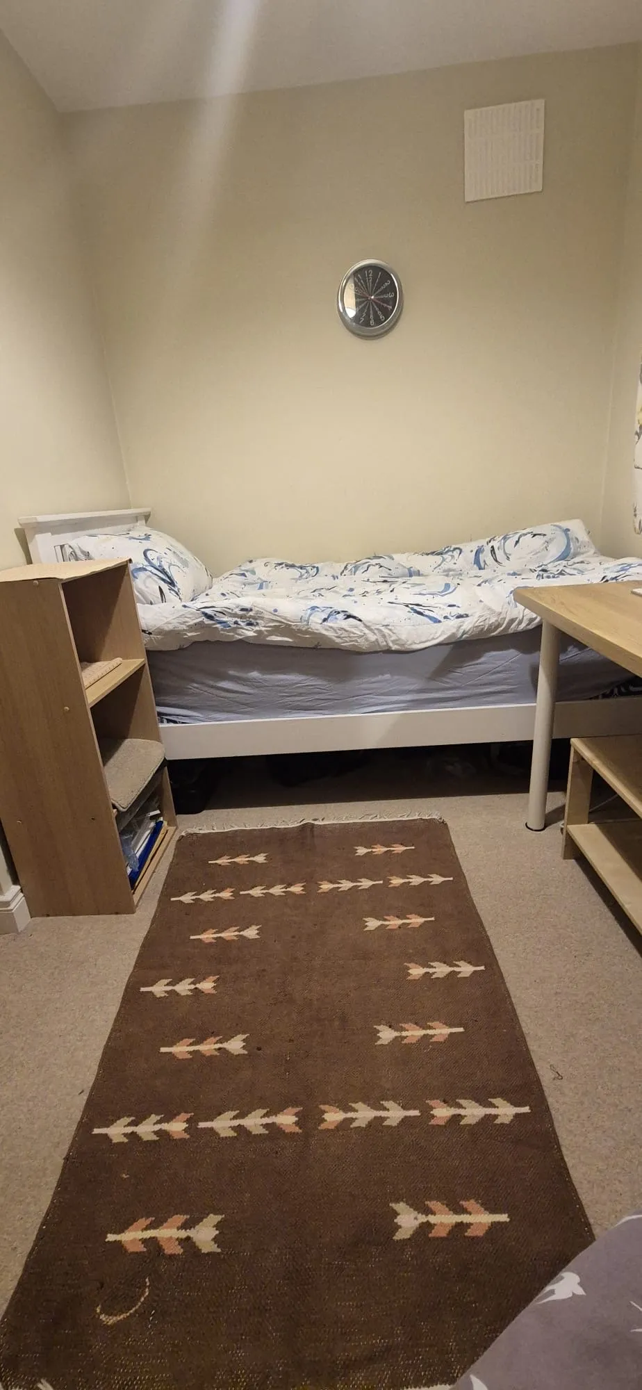 Small bedroom with a single bed against a wall, patterned duvet, wall clock above the bed, wooden shelving unit at the foot, a desk to the right, and a brown patterned runner rug on the floor.