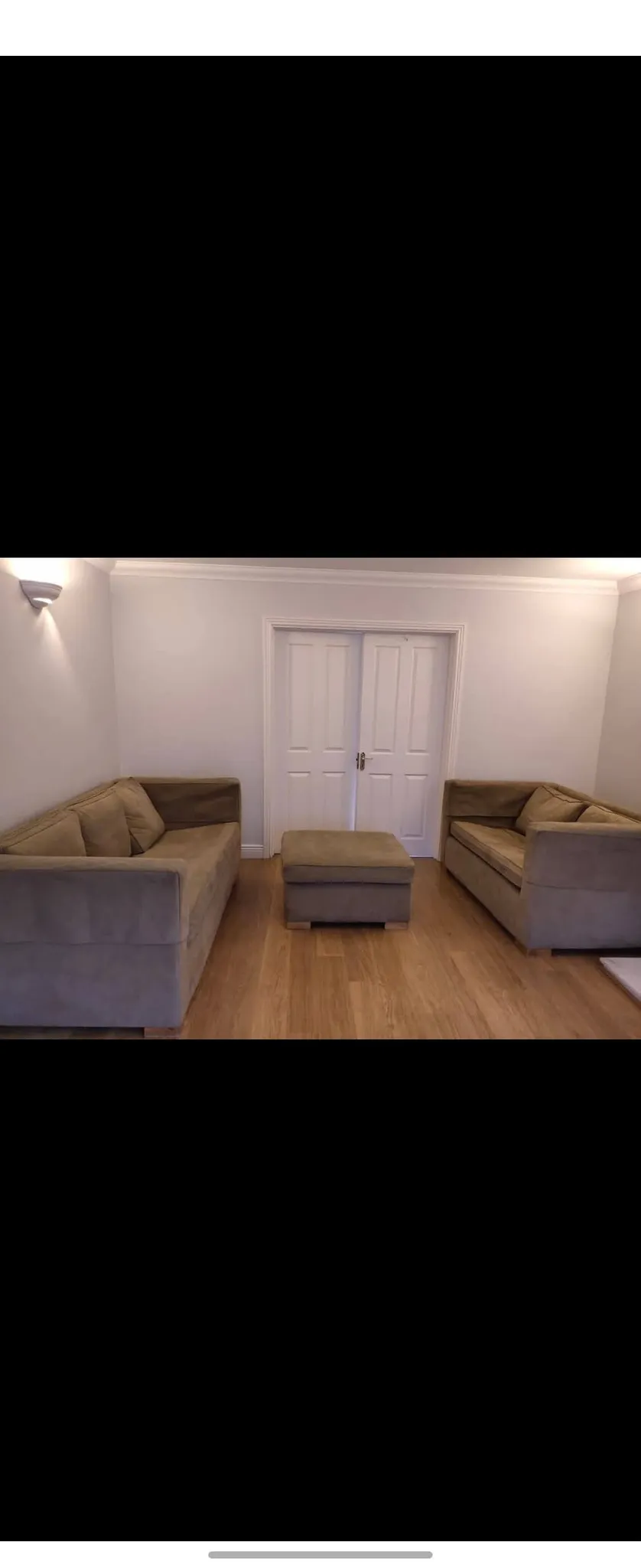 Living room with two sofas facing each other, a central ottoman, wooden floor and closed double doors on the far wall.