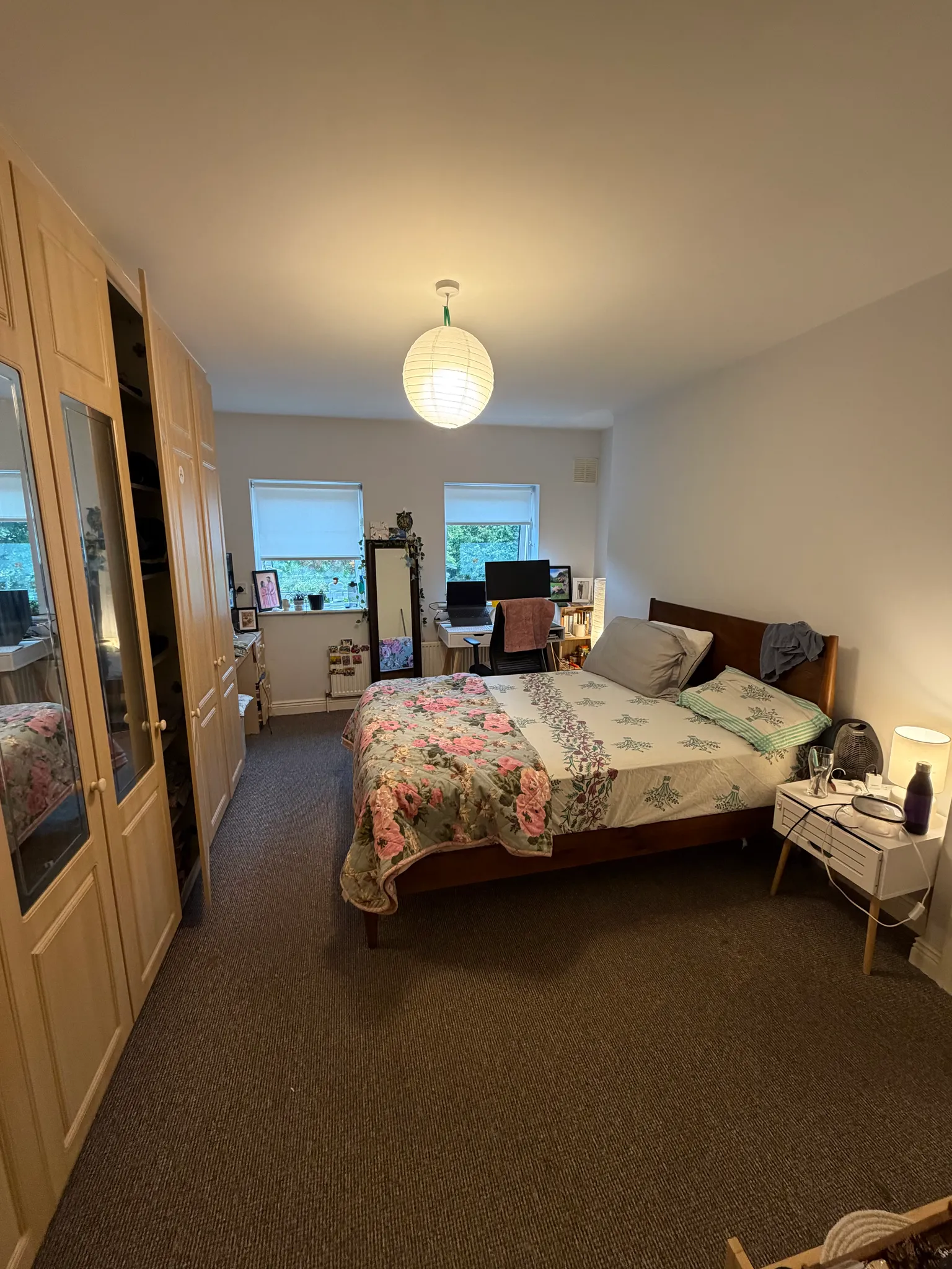Bedroom with a double bed, bedside table and lamp, wardrobe along the left wall, desk and computer under two windows, and a hanging ceiling light.