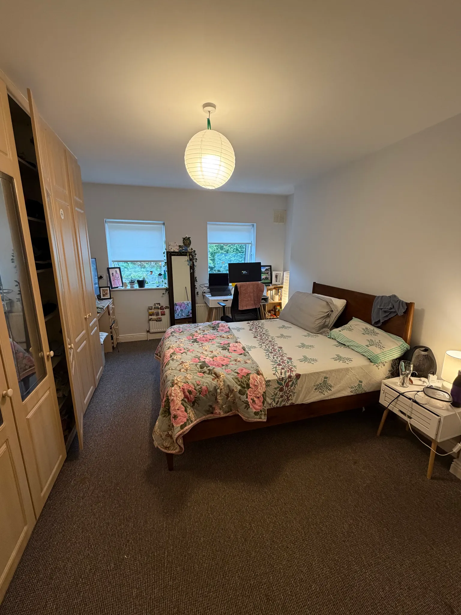 Bedroom with a double bed, bedside table, wardrobe, desk with a computer, two windows and a hanging ceiling light.