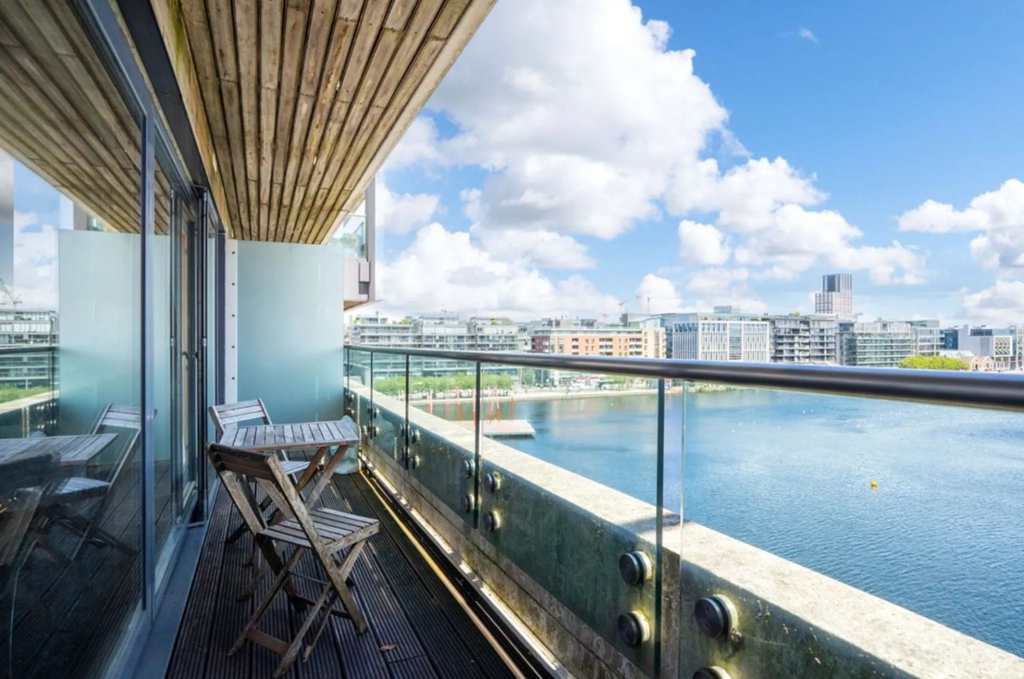 Balcony with a small wooden table and folding chairs, glass railing and view over water and nearby buildings.